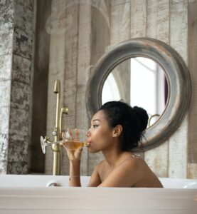 Side view of young ethnic female sitting in bathtub and drinking wine while enjoying spa procedure in bathroom